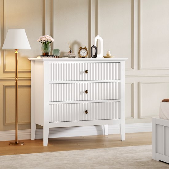 Commode blanche à trois tiroirs nervurés et boutons dorés, à côté d'une lampe sur pied.