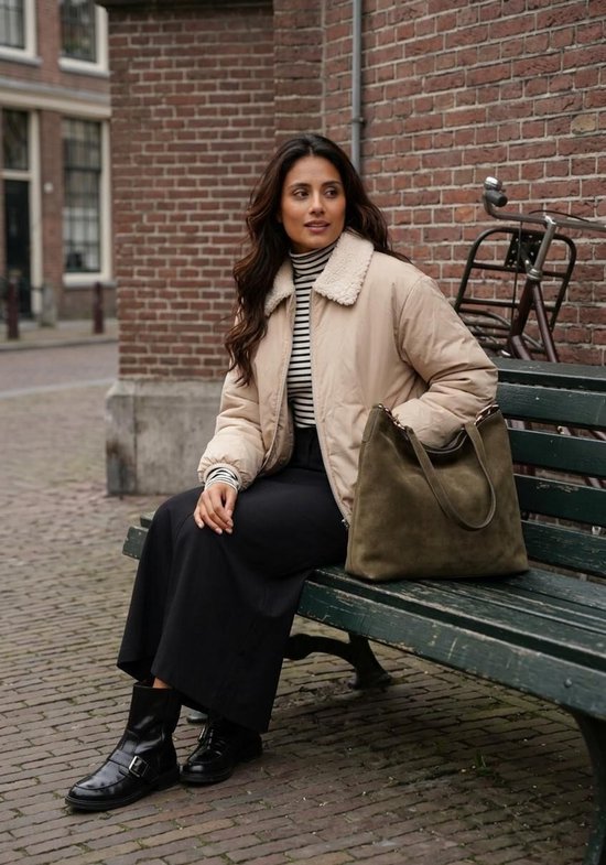 Vrouw op een bank met beige teddy-jas, gestreepte coltrui, zwarte rok, zwarte laarzen en een olijfgroene suède tas.