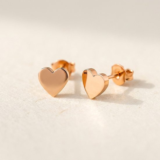 Rosegouden hartvormige oorknopjes op een zachte beige ondergrond.