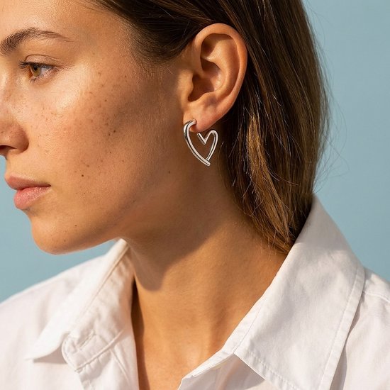 Model draagt zilverkleurige hartvormige oorbellen met een wit overhemd.