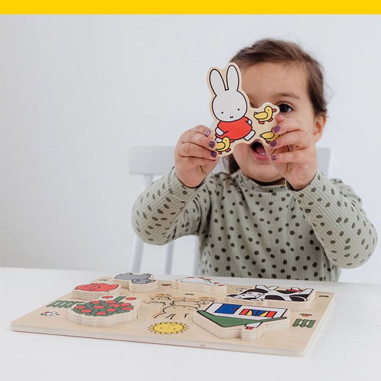 Een meisje met bruine krulletjes houdt een Miffy-puzzelstukje met kuikentjes omhoog, met een boerderijpuzzel op tafel.