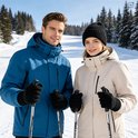 Man in blauwe en vrouw in beige jas, beiden dragen zwarte gebreide handschoenen en skiën in een besneeuwd landschap.