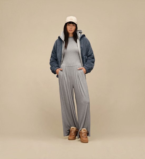 Model draagt een blauw sherpa jack, grijze jumpsuit, beige UGG-pet en bruine suède Lowmel-sneakers.
