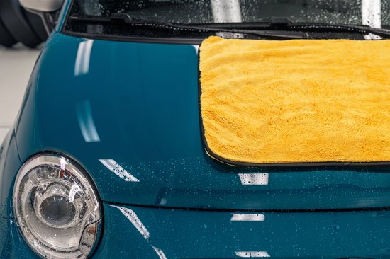 Een felgeleende microvezel droogdoek op de teal blauwe motorkap van een auto, met waterdruppels en koplamp.
