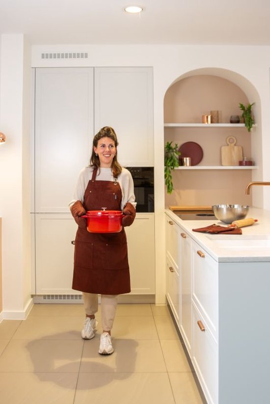 Vrouw in een bruin schort en bijpassende Tiseco ovenwanten draagt een rode braadpan in een moderne keuken.