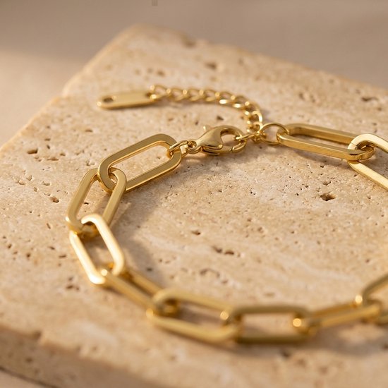 Gouden schakelarmband op een beige textuursteen, met focus op de karabijnsluiting.