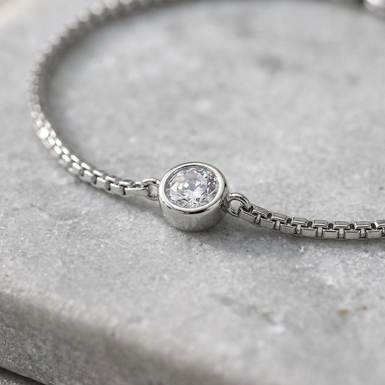 Close-up van een zilveren armband met een centrale ronde zirkonia steen op een grijze stenen ondergrond.