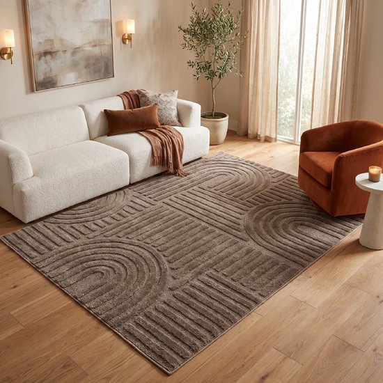 Modern bruin vloerkleed met geometrisch patchworkpatroon, in een lichte woonkamer met witte bank en oranje fauteuil.
