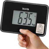 Hand die de compacte, zwarte Tanita weegschaal vasthoudt, met een 'Reisformaat' sticker.