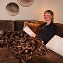 Vrouw met AYO-lichttherapiebril die rood licht uitstraalt, ontspant op een bank onder een zachte bruine deken.