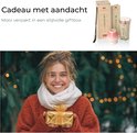Vrouw met witte sjaal en roestkleurige trui houdt een gouden cadeau vast voor een verlichte kerstboom.