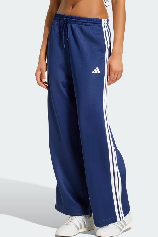 Model draagt een wijde donkerblauwe adidas trainingsbroek met wit logo, trekkoord en witte strepen op de broekspijpen.