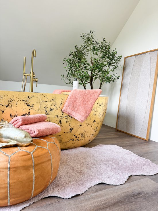 Luxe badkamer met goudkleurig bad, zalmroze handdoeken, een poef, een plant en een lichtroze badmat.
