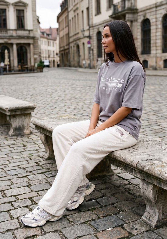 Grijze New Balance oversized t-shirt met 'New Balance Athletics' logo op de borst, gedragen door een vrouw zittend op een stenen bankje.