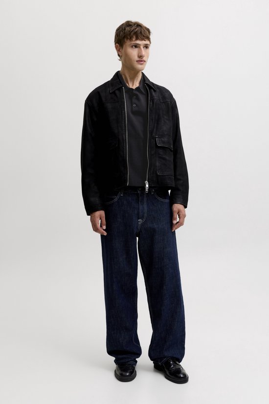 Model draagt een zwarte bomberjack over een zwart poloshirt en donkere wijde jeans.