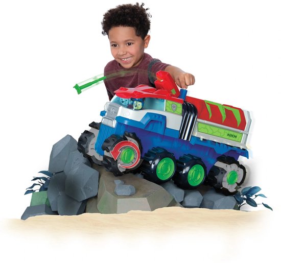 Een jongen speelt met een blauwe PAW Patrol vrachtwagen op een rotsachtige ondergrond, terwijl hij een groen projectiel lanceert.