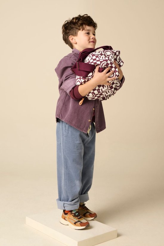 Jongen met paars corduroy overhemd, blauwe corduroy broek en tas met repetitief patroon.