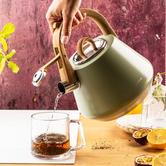 Hand schenkt water uit een matgroene Klausberg ketel met houtlook handvat in een glazen theekop.