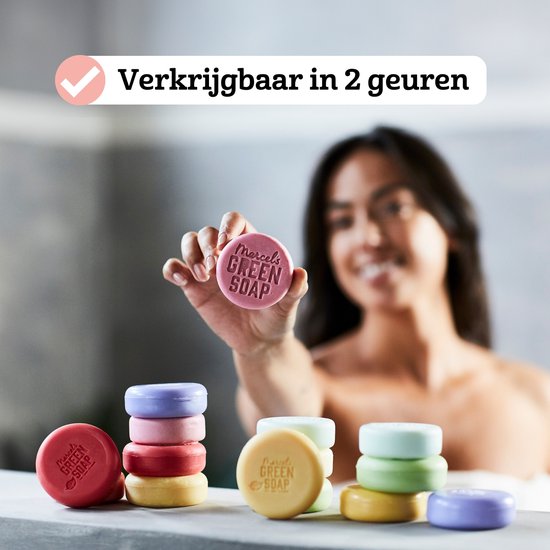 Een vrouw houdt een roze shampoo bar vast. Stapels gekleurde shampoo bars liggen op een toonbank.