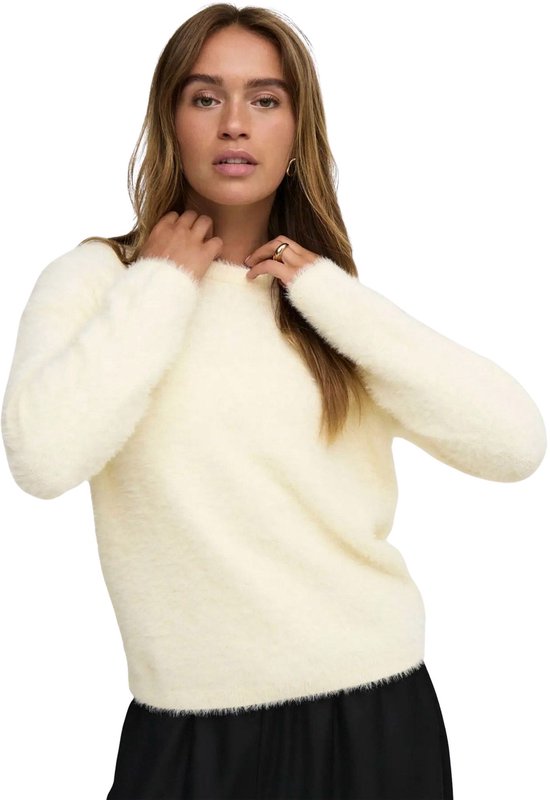 Model in crèmekleurige, pluizige trui, handen bij de hals, zwarte broek.