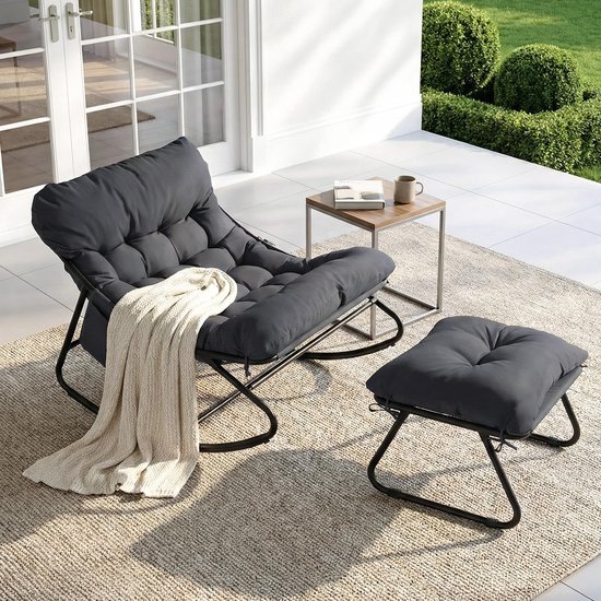 Grijze schommelstoel met voetenbank, gedrapeerd met een beige plaid, op een terras bij zonnig weer.