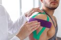 Een schouder van een man met groene en paarse sporttape, aangebracht door een persoon in een witte doktersjas.