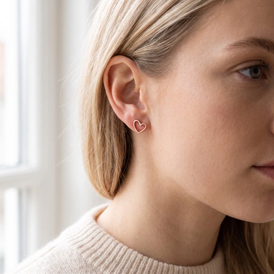 Close-up van een vrouw met blond haar, die een rosegoudkleurige hartjesoorbel met opengewerkte vorm draagt in haar linkeroor.