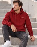 Man met donkere jeans, witte sneakers en rode COVAR hoodie met capuchon, zittend.
