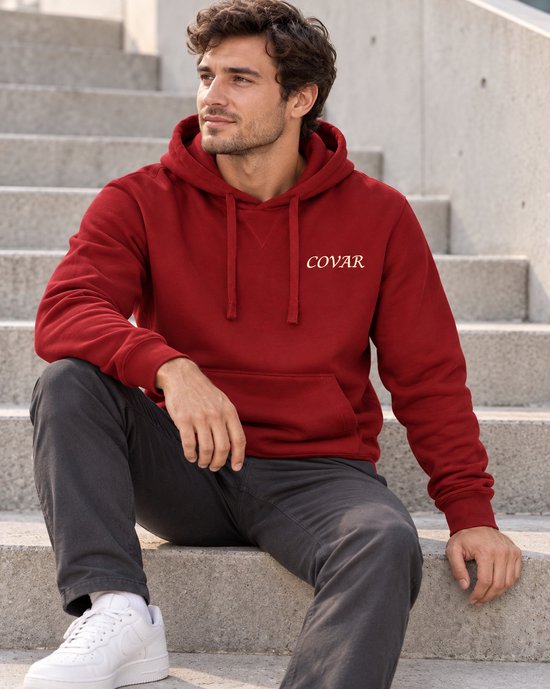 Man met donkere jeans, witte sneakers en rode COVAR hoodie met capuchon, zittend.