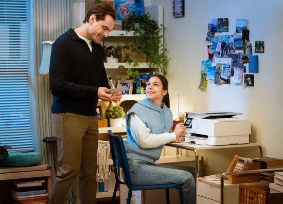 Man en vrouw in thuiskantoor; vrouw bedient witte HP Envy Photo printer terwijl man haar een document overhandigt.