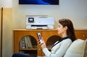 Vrouw met beige vest houdt smartphone vast voor een witte HP Envy Photo printer op een houten dressoir.