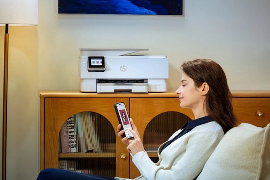 Vrouw met beige vest houdt smartphone vast voor een witte HP Envy Photo printer op een houten dressoir.