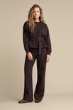 Model met lang blond golvend haar draagt een donkerbruine sweater met ceintuur en wijde broek.