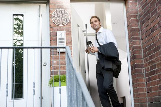 Man met een grijze/zwarte reismok en een jas over zijn schouder, verlaat een huis.