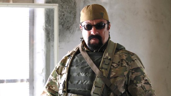 Man met snor en baard draagt een camouflagepet, zonnebril, tactisch vest en camouflagejas.
