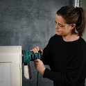 Vrouw met veiligheidsbril gebruikt groene en zwarte excentrische schuurmachine op een wit meubel.