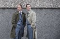 Twee mannen in trenchcoats en pakken staan tegen een grijze, ruw gestructureerde muur.