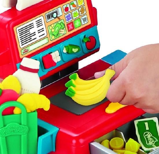 Een hand legt een gele Play-Doh banaan op een rode en blauwe speelgoedkassa met een groene boodschappentas.
