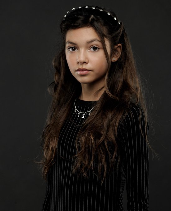 Close-up van een meisje met lang bruin haar, dat een zwarte jurk met witte strepen en een ketting met letters draagt.