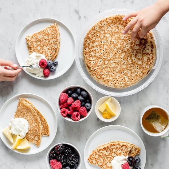 Table avec des crêpes, des fruits, du citron et une tasse de thé. Quelqu'un prend une crêpe dans la boîte de conservation.