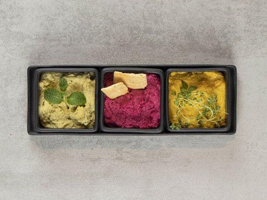 Zwarte serveerschaal met drie vakken gevuld met dips en crackers. Groene dip met munt, rode dip met crackers, gele dip met kruiden.