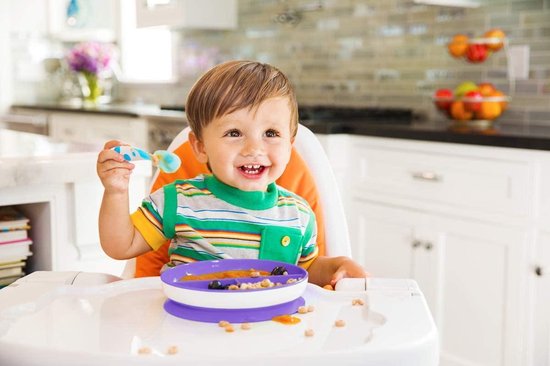 Bébé joyeux dans une chaise haute mangeant avec une cuillère bleue et blanche à pois