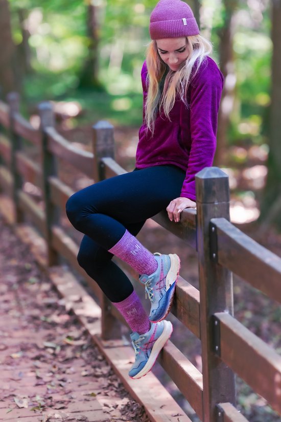 Vrouw met blonde haren in paarse muts en trui, zwarte legging, roze sokken en blauw-roze wandelschoenen, zittend op een houten hek in het bos.