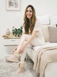 Een lachende vrouw in een witte top en crème broek, zittend op een bed, draagt beige pantoffels.