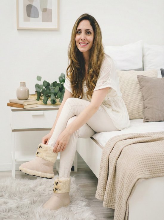 Een lachende vrouw in een witte top en crème broek, zittend op een bed, draagt beige pantoffels.
