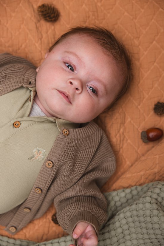 Close-up van baby met geborduurd boompje op lichtgroene romper, bruin vestje met knopen, liggend op oranje plaid.