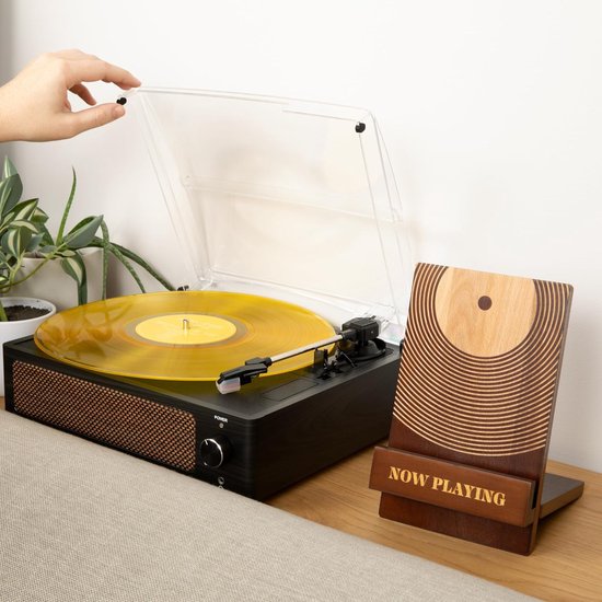 Hand plaatst gele vinylplaat op platenspeler, met houten "NOW PLAYING"-standaard ernaast.