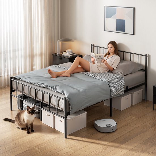 Une femme lit un livre dans un lit avec un cadre en métal noir, une couette grise, une couverture à carreaux et des boîtes de rangement sous le lit. Un chat est assis à côté.