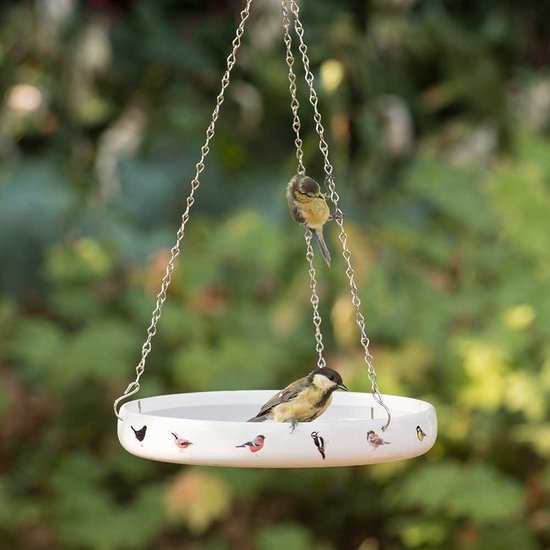 Wit porseleinen vogelvoederschaaltje met kleine vogels erop afgebeeld, hangend aan een ketting. Twee koolmezen zitten aan de rand en op de ketting.
