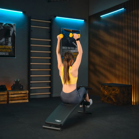 Vrouw met gele halter heft boven het hoofd, zittend op de schuine trainingsbank.